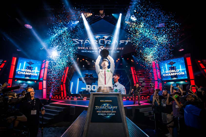 - 香港Gameone娛樂城全新會員福利 - 入金00即返送00! Esports player in a white jacket raises a trophy on a brightly lit stage with blue confetti during the StarCraft World Championship Series finals, crowd cheering in the foreground.