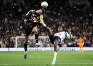 Two soccer players leap in the air competing for a header during a match, one in a dark kit with a Red Bull logo and the other in a white jersey marked USA on the chest.
