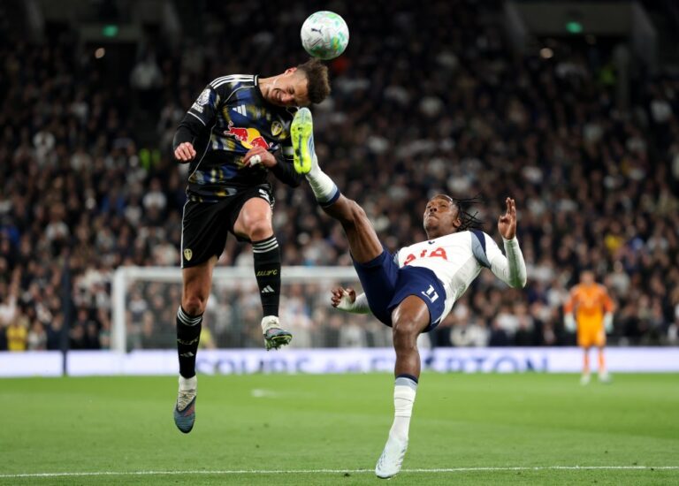 Two soccer players leap in the air competing for a header during a match, one in a dark kit with a Red Bull logo and the other in a white jersey marked USA on the chest.