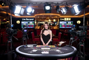 - 香港Gameone娛樂城全新會員福利 - 入金00即返送00! - 香港Gameone娛樂城全新會員福利 - 入金00即返送00! Hostess in a black dress stands at a casino baccarat table, surrounded by cameras and bright studio lights in a live‑casino setup.