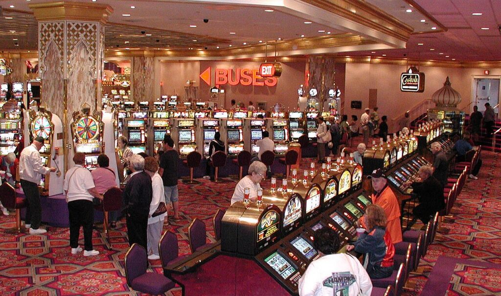 - 香港Gameone娛樂城全新會員福利 - 入金00即返送00! Busy casino scene with rows of slot machines and players seated or standing at them in a colorful, carpeted hall.