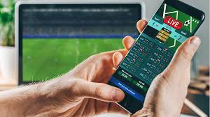 Person holds a smartphone showing live sports betting odds while a laptop screen in the background displays a stadium game.