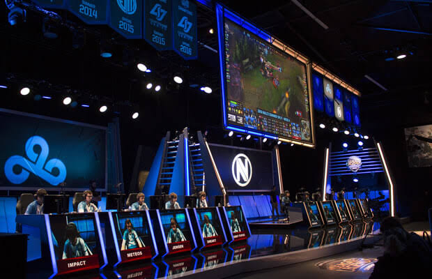 Esports tournament stage with players at neon-lit desks and a large screen showing gameplay.